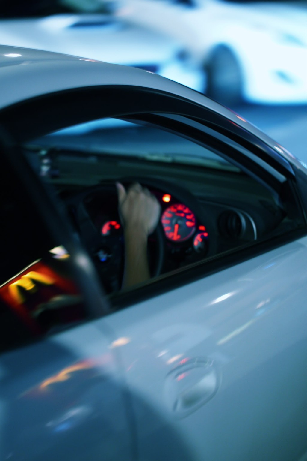 A driver’s hand and red dashboard lights framed by the window of a white car