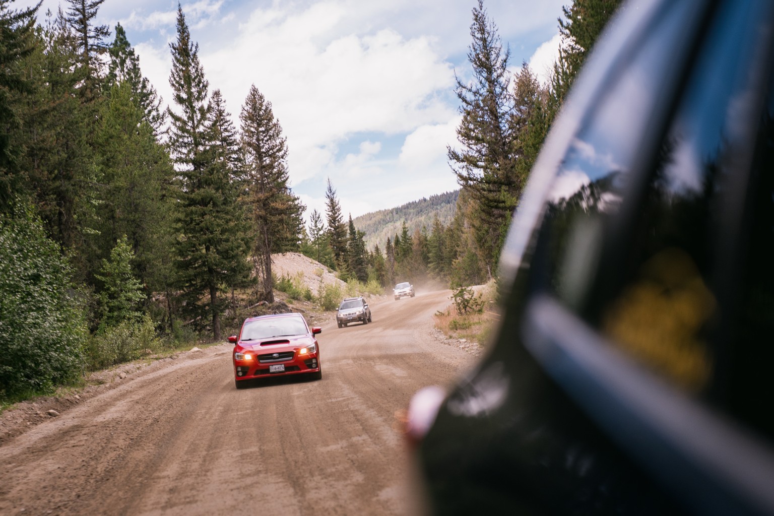 A convoy of cars following a gravel road through a forest, viewed from a moving vehicle