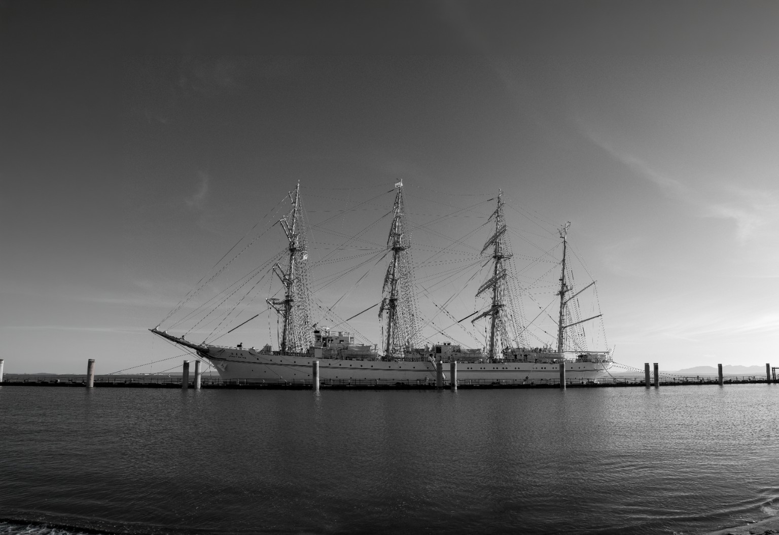 Black-and-white photograph of a four-masted sailing ship moored beside calm water